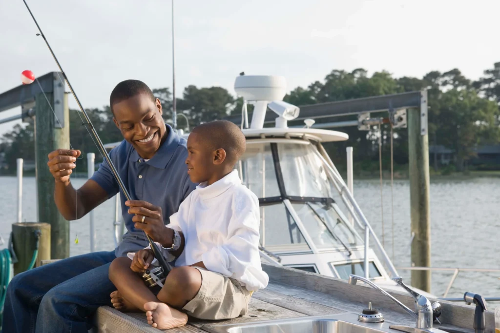 A man and a boy fishing together on a small boat, surrounded by calm water and a clear blue sky.